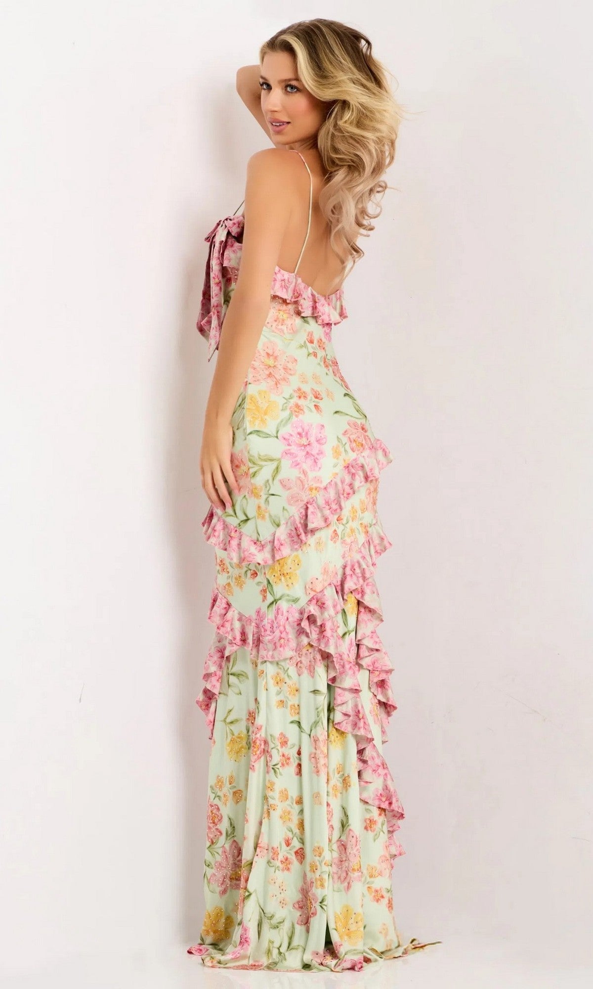 A woman models the Jovani 47236 Ruffled Long Floral-Print Prom Gown, featuring thin straps and an open back, as she faces away from the camera against a plain light background.